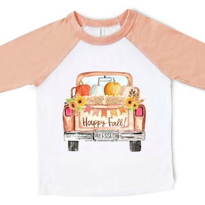 May include: A white and peach long-sleeved t-shirt with a watercolor illustration of a brown pickup truck carrying pumpkins, hay bales, and sunflowers. The truck has a license plate that says "BLESSED" and the text "Happy Fall!" is printed on the back.