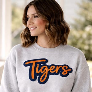May include: A light gray sweatshirt with the word "Tigers" in orange script lettering outlined in navy blue. The sweatshirt is a crew neck style.