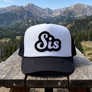 May include: A black and white trucker hat with the word "Sis" in a cursive font on the front. The hat has a white front panel, a black brim, and a black mesh back. The hat is sitting on a wooden picnic table with a mountain range in the background.