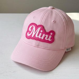 May include: A light pink baseball cap with a curved brim. The cap features a pink patch with the word "Mini" in white script lettering. The cap is designed for ages 3-6.