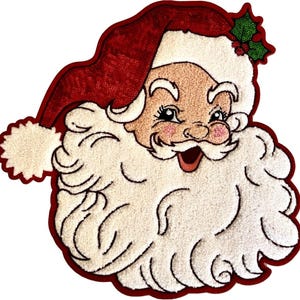 May include: A Santa Claus head patch featuring a red sequined hat with a white pom-pom, a thick white textured beard, and a green holly leaf accent with red berries. The design has a dark red border and a light peach-colored face.