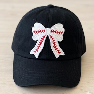 Peut inclure: Casquette de baseball noire avec un écusson blanc en forme de nœud. Le nœud présente des coutures rouges, ressemblant à une balle de baseball. La casquette a une visière incurvée et un bouton sur le dessus. Un accessoire élégant.