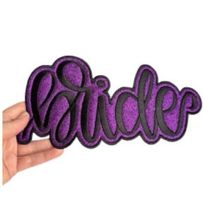 May include: A purple glitter patch with the word "Bride" in black script lettering. The patch is held in a hand against a white background. The patch is a decorative item.