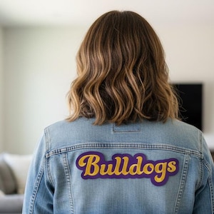 May include: A light blue denim jacket with a purple and gold "Bulldogs" patch on the back. The jacket is worn by a person with wavy brown hair. The background is a blurred interior with a couch and television.