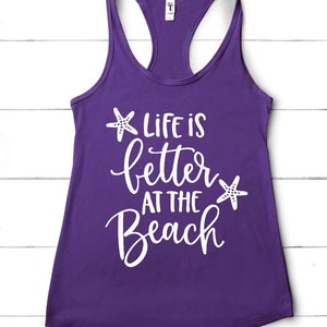 Pu&ograve; includere: Canotta viola con testo bianco che recita "Life is better at the beach" con una stella marina bianca su entrambi i lati del testo.