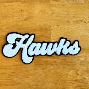 May include: White felt "Hawks" lettering with a black outline. The letters are in a retro, cursive style.
