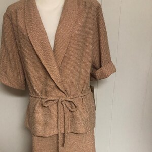 May include: A two-piece skirt suit in a light brown color with a subtle shimmer. The jacket has a shawl collar and a tie belt. The skirt is knee-length.
