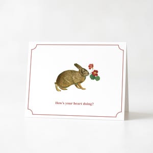 Peut inclure: Carte de vœux blanche avec une bordure rouge et le texte "How's your heart doing ?" présente un lapin brun tenant des fleurs rouges. La carte a un design simple.