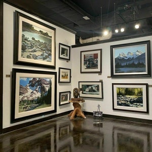 May include: An art gallery featuring framed landscape paintings and prints. The artwork depicts mountains, rivers, and forests. A canoe is mounted on the wall. Postcards are displayed on a rotating rack.