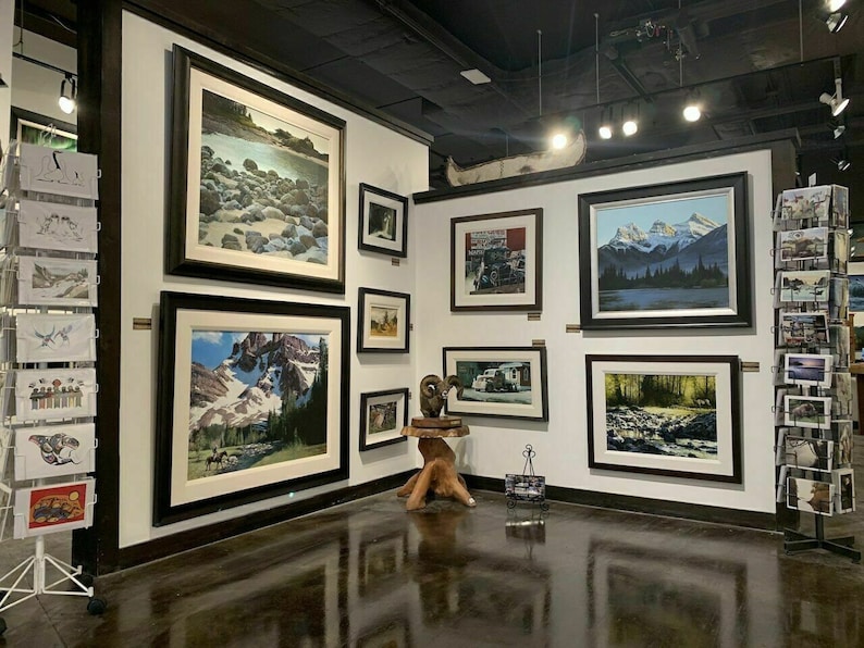 May include: An art gallery featuring framed landscape paintings and prints. The artwork depicts mountains, rivers, and forests. A canoe is mounted on the wall. Postcards are displayed on a rotating rack.