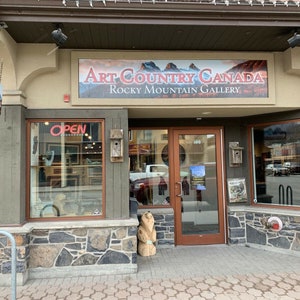 May include: Exterior view of the Art Country Canada Rocky Mountain Gallery. The storefront features a large sign with the gallery's name and a mountain landscape. A smaller sign hangs to the left. The entrance has a glass door with the number 100. An "OPEN" sign is visible.