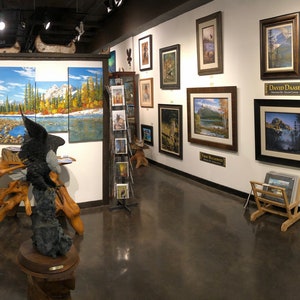 May include: An art gallery interior featuring framed landscape paintings and sculptures. The walls are adorned with various artworks, including paintings of mountains, rivers, and sunsets. A glass display case showcases additional items. The gallery has a polished concrete floor.