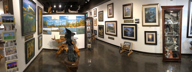 May include: An art gallery interior featuring framed landscape paintings and sculptures. The walls are adorned with various artworks, including paintings of mountains, rivers, and sunsets. A glass display case showcases additional items. The gallery has a polished concrete floor.