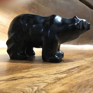 Puede incluir: Una figura de oso negro con un acabado liso y brillante. El oso se representa de pie, con sus cuatro patas firmemente plantadas. La escultura es de un color oscuro sólido, y los detalles de la forma del oso son claramente visibles.