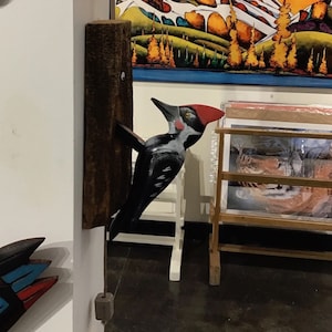 May include: A wooden woodpecker sculpture, painted black, white, and red, is mounted on a dark brown wooden post. The bird has a red crest and is positioned as if pecking at the wood. A small weight hangs below.