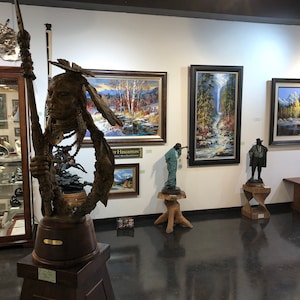 May include: An art gallery interior featuring various sculptures and framed paintings. A large bronze sculpture of a Native American figure holding a spear is prominent. Paintings depict landscapes, and other sculptures are displayed on pedestals and tables.