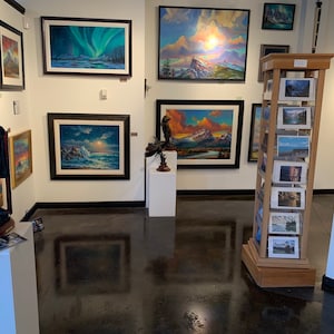 May include: An art gallery interior featuring framed landscape paintings with various scenes, including mountains, sunsets, and aurora borealis. A wooden display rack holds smaller landscape prints. A bronze statue of a man stands on a white pedestal.