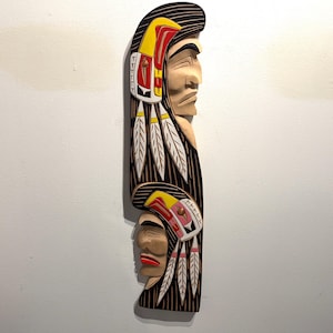 Puede incluir: Un colgante de pared de madera tallada que representa dos figuras nativas americanas de perfil. Las figuras tienen tocados detallados en rojo, amarillo y blanco. La talla tiene un fondo oscuro con líneas verticales y está montada en una pared blanca.