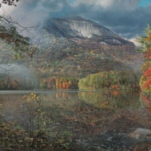 May include: A scenic landscape painting of a lake surrounded by trees in autumn colors. The water reflects the trees and a mountain in the background. The sky is overcast with clouds, creating a moody atmosphere.