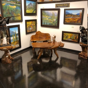 May include: An art gallery featuring framed landscape paintings with mountain and water scenes. The paintings are displayed on white walls, with a wooden bench and table in the center. A wooden sculpture of a person is on the left.