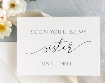 May include: A white card with black text that reads "Soon you'll be my sister. Until then..." The card is surrounded by pink and white flowers and a light pink ribbon.