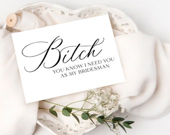 May include: A white card with black cursive text that reads "Bitch, you know I need you as my bridesmaid." The card is placed on a white, textured surface with a sprig of greenery and white flowers.