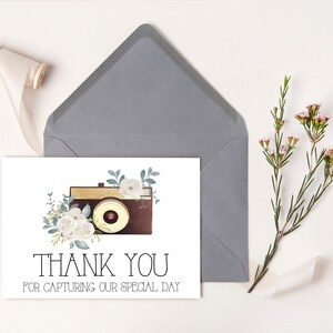 May include: A white thank you card with a vintage camera illustration surrounded by white flowers and green leaves. The text on the card reads "Thank You for Capturing Our Special Day."