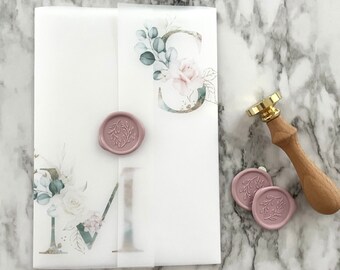 Puede incluir: Un sobre blanco con un diseño floral y un sello de cera rosa. El sobre está sobre una superficie de mármol con un sello de cera dorado y de madera.
