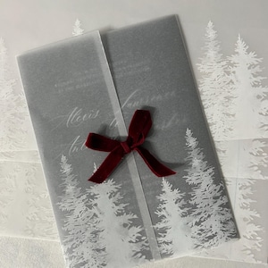 May include: A gray invitation wrapped in translucent paper with a white pine tree design. The invitation is tied with a dark red velvet ribbon. The text "Alexis" is visible on the invitation.