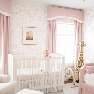 May include: A white wooden crib with a ruffled skirt and patterned sheet, positioned in a room with pink floral wallpaper, pink curtains with white valances, a framed art print, a plush giraffe toy, and a pink upholstered daybed.