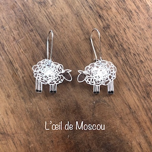 Peut inclure: Une paire de boucles d'oreilles pendantes argentées avec un motif de mouton blanc. Les moutons sont soulignés de noir et ont un motif de laine bouclée.