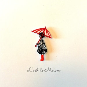 May include: A black and white patterned paper doll with a red umbrella and red boots. The doll is holding the umbrella and is standing on a white background. The text "L'oeil de Moscou" is written below the doll.