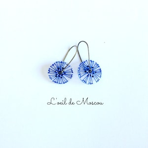 Puede incluir: Un par de pendientes de flores azules y blancas con ganchos plateados. Los pendientes están hechos de resina y tienen un diseño delicado pintado a mano.