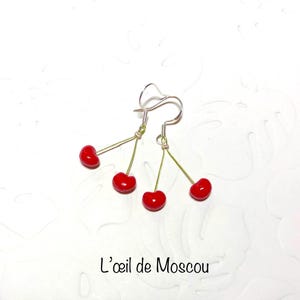 Peut inclure: Boucles d'oreilles pendantes argentées avec trois breloques cerises rouges. Les boucles d'oreilles sont sur un fond blanc.