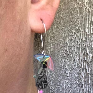 K&ouml;nnte beinhalten: Silberner Creolen-Ohrring mit einem Anh&auml;nger, der eine Person mit einem bunten Regenschirm zeigt. Die Person tr&auml;gt ein schwarzes Kleid mit geometrischem Muster und rosa Stiefel. Der Regenschirm hat ein Regenbogen-Design.