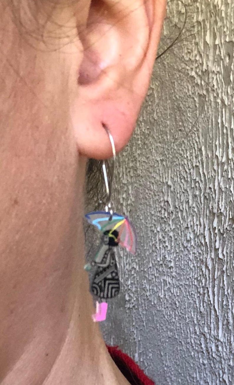K&ouml;nnte beinhalten: Silberner Creolen-Ohrring mit einem Anh&auml;nger, der eine Person mit einem bunten Regenschirm zeigt. Die Person tr&auml;gt ein schwarzes Kleid mit geometrischem Muster und rosa Stiefel. Der Regenschirm hat ein Regenbogen-Design.