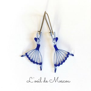 Puede incluir: Un par de pendientes de bailarina azul y blanco con ganchos dorados. Las bailarinas están en una pose de baile con los brazos levantados por encima de sus cabezas. Los pendientes están hechos de resina y son ligeros y cómodos de llevar.