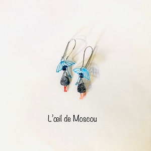 Puede incluir: Un par de pendientes de plata con figuras de patrones azules y negros que sostienen paraguas azules. Los pendientes están etiquetados como "L'oeil de Moscou".