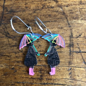 May include: A pair of dangle earrings featuring a design of two figures under colorful umbrellas. The figures are wearing black dresses and pink boots. The earrings have silver-colored hooks and are set against a wooden background.