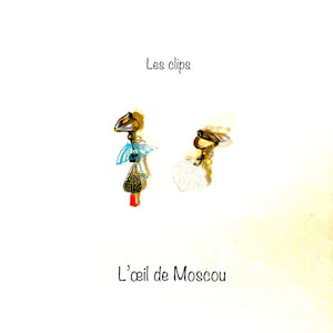 Puede incluir: Un par de pendientes de clip con un diseño estampado en azul y blanco con un acento rojo. Los pendientes están etiquetados como "Les clips" y "L'oeil de Moscou".