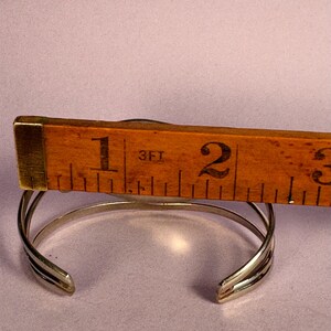 May include: A silver cuff bracelet with a layered design, shown with a wooden ruler measuring its width. The bracelet is approximately 1 inch wide. The background is a solid light purple color.