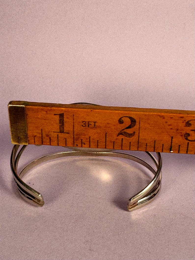 May include: A silver cuff bracelet with a layered design, shown with a wooden ruler measuring its width. The bracelet is approximately 1 inch wide. The background is a solid light purple color.