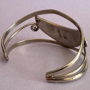 May include: A silver-toned cuff bracelet with an open design. The bracelet features a central, leaf-shaped element and a decorative swirl. The word "MEXICO" is stamped on the inside of the leaf-shaped element.