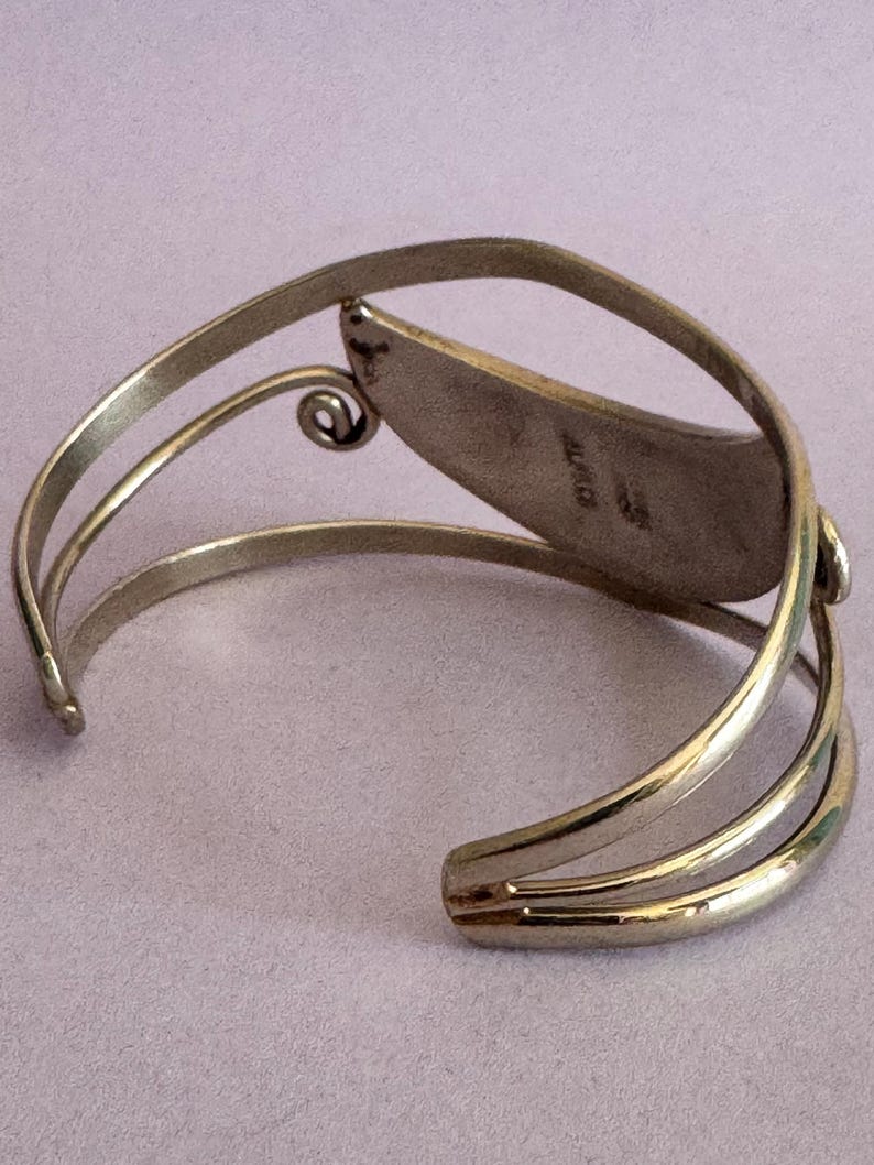 May include: A silver-toned cuff bracelet with an open design. The bracelet features a central, leaf-shaped element and a decorative swirl. The word "MEXICO" is stamped on the inside of the leaf-shaped element.