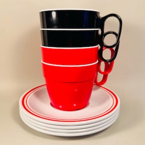 May include: A stack of four coffee cups in black and red, with white rims, and a stack of matching saucers. The cups have loop handles. The saucers are white with a red stripe.