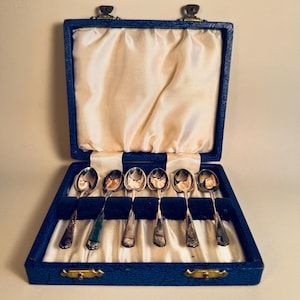 Op de afbeelding: Een set van zeven zilveren lepels gepresenteerd in een blauwe fluwelen doos met een crèmekleurig satijnen interieur. De lepels hebben een klassiek ontwerp en de doos is afgesloten met een sluiting.