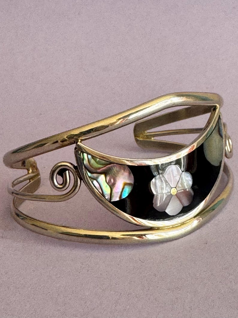 May include: A gold-toned cuff bracelet with a black inlay. The inlay features an abalone shell design and a mother-of-pearl flower. The bracelet has a curved design with a decorative swirl element.