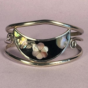 May include: Silver-tone cuff bracelet with a black inlay featuring a flower design. The flower is made of mother-of-pearl, with iridescent accents. The bracelet has a curved design with decorative swirls on each side.