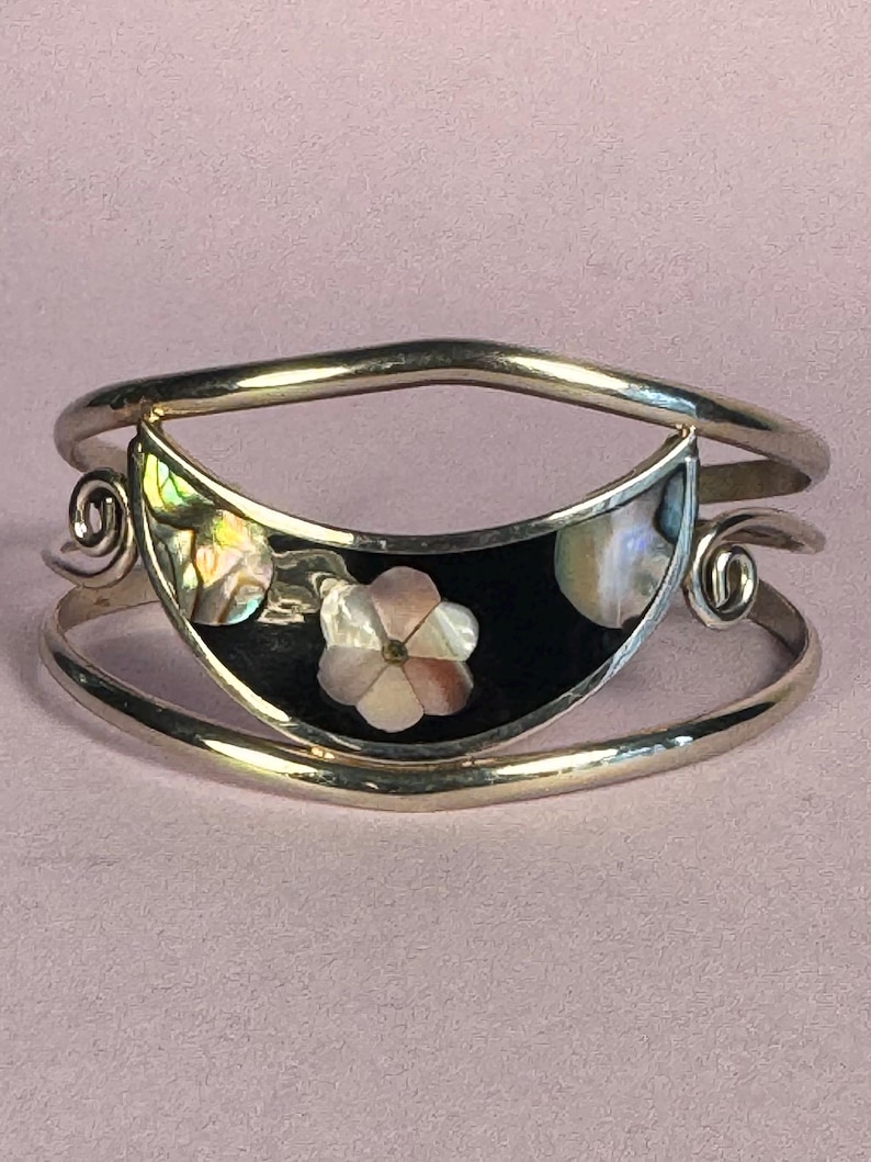 May include: Silver-tone cuff bracelet with a black inlay featuring a flower design. The flower is made of mother-of-pearl, with iridescent accents. The bracelet has a curved design with decorative swirls on each side.