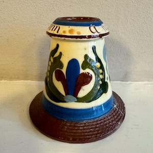 Puede incluir: Un portavelas de cerámica pintado a mano con un cuerpo color crema, adornado con diseños florales azules, verdes y granates. La base es un anillo texturizado marrón oscuro, y la parte superior presenta una banda azul y detalles decorativos.
